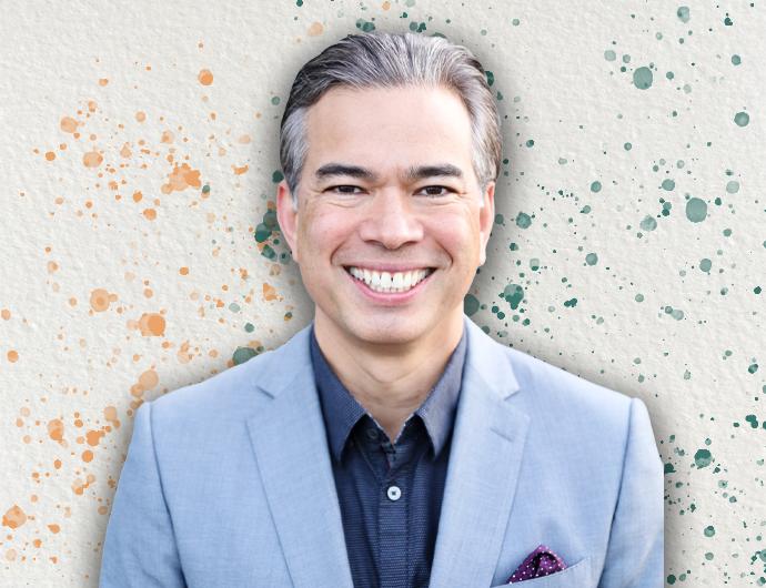 Headshot of a smiling Rob Bonta, in a light blue-grey suit, on top of an off-white background with orange and green 