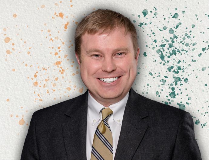 Headshot of AG Aaron Frey in a black suit with gold striped tie, over an off-white background with orange and green spatter