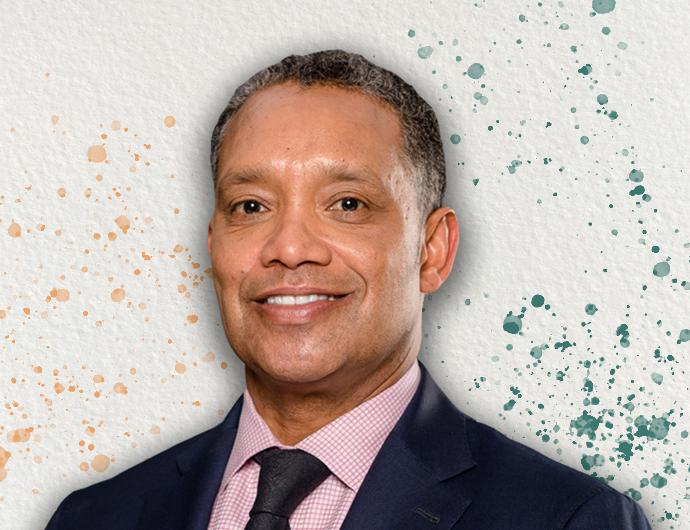 Headshot of AG Karl Racine in a navy suit with black tie, over an off-white background with orange and green spatter